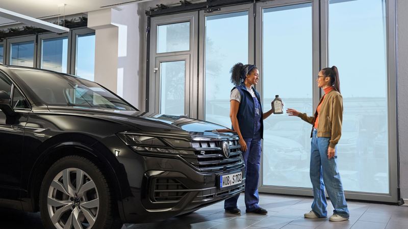 VW Servicemitarbeitender übergibt dem Kunden eine Flasche Motorenöl neben einem VW Touareg