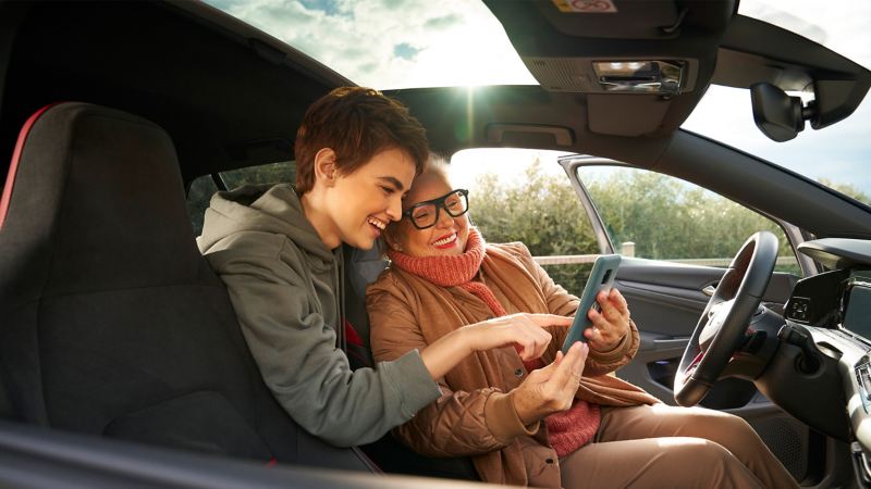 Twee vrouwen in een VW met geopende bestuurdersdeur kijken naar een smartphone – Entertainment en elektronica