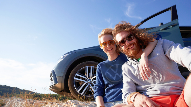 Frau und Mann in stylischer Kleidung sitzen vor VW Auto – VW Fanartikel