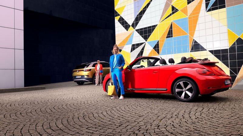 Δύο νέες γυναίκες στα αυτοκίνητά τους VW, ID.4 και Beetle Cabriolet, μπροστά σε έναν πολύχρωμο τοίχο – Ιδιοκτήτες και υπηρεσίες After Sales VW