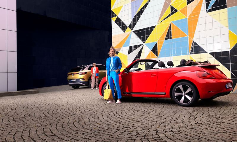 Two young women at their VW cars, ID.4 and Beetle Cabriolet, in front of a colourful wall – owners and services