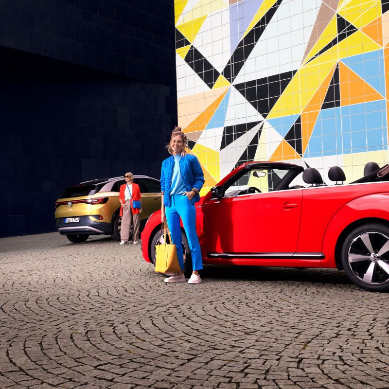 Two young women at their VW cars, ID.4 and Beetle Cabriolet, in front of a colourful wall – owners and services