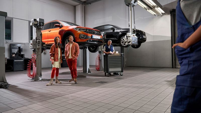 Twee klanten en een VW-servicemedewerker in de werkplaats, VW T-Cross en VW Touareg op liftplatforms