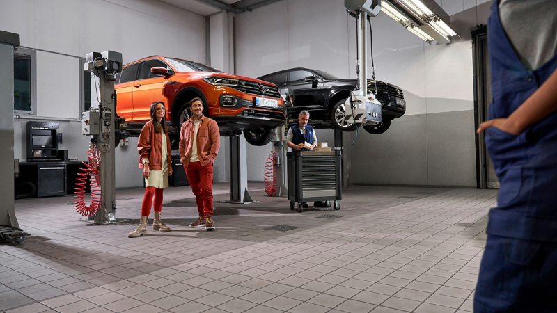 Dos clientes y un empleado del servicio técnico de VW en el taller, VW T-Cross y VW Touareg sobre plataformas elevadoras