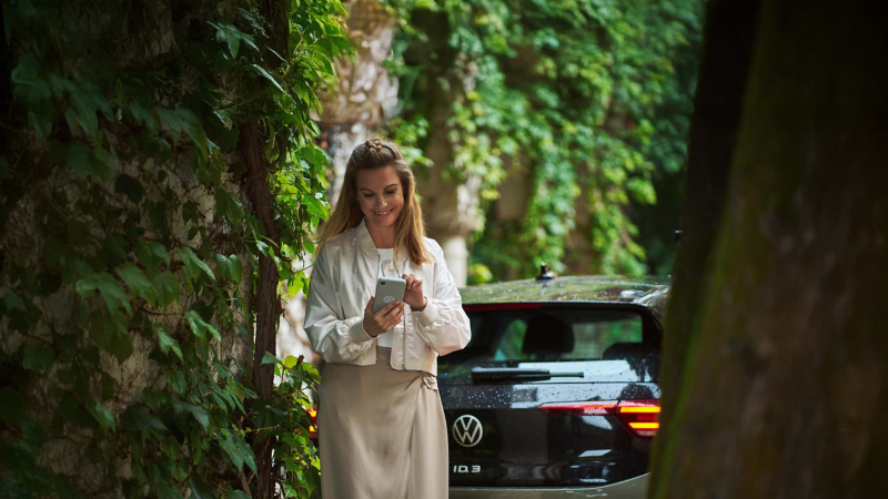 Eine Frau steht vor ihrem Volkswagen und schaut auf ihr Smartphone