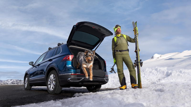 A man picks up a snowboard from the ski and snowboard holder of his silver VW car with winter tyres in a snowy landscape