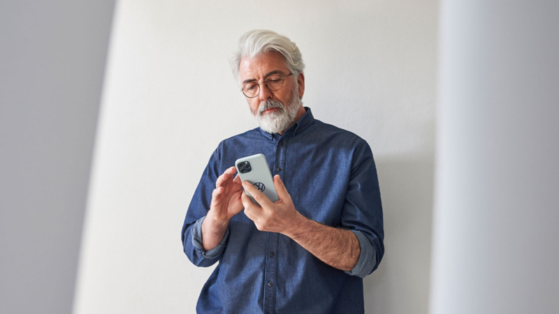 Mann mit grauem Haar und Bart steht vor einer hellen Wand und schaut konzentriert auf sein Handy.
