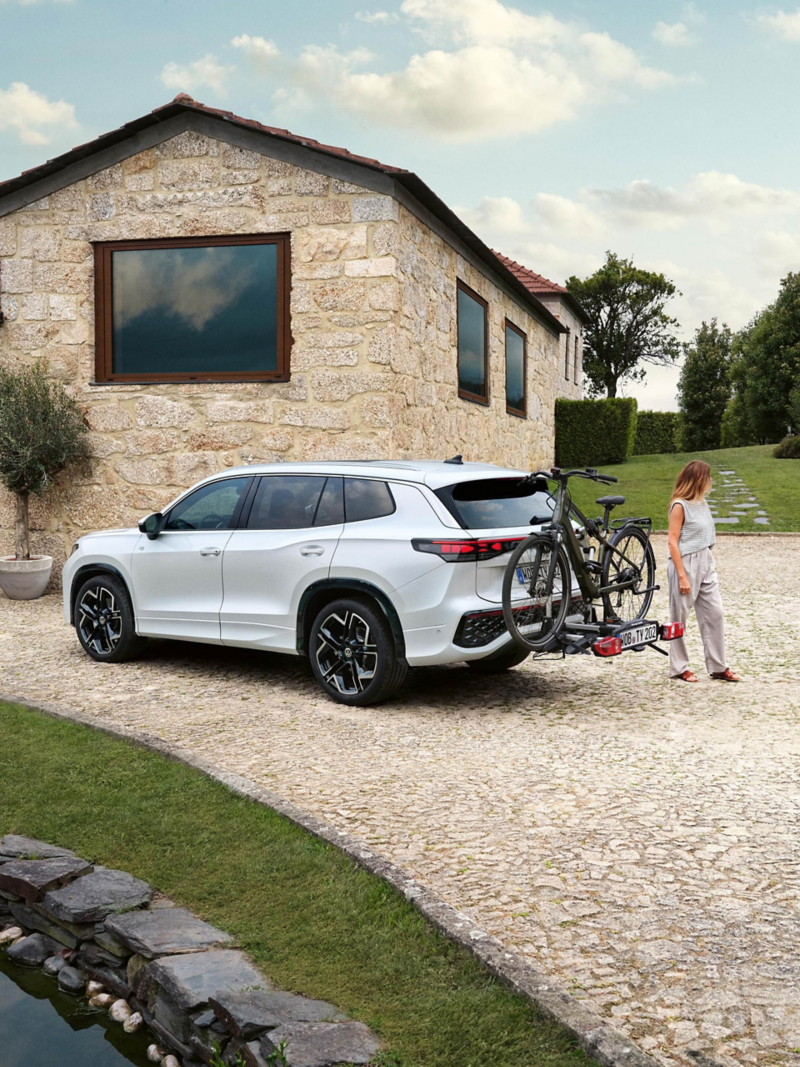 Una mujer de pie junto a un Volkswagen Tayron con una bicicleta en el portabicicletas, llega un hombre empujando una bicicleta