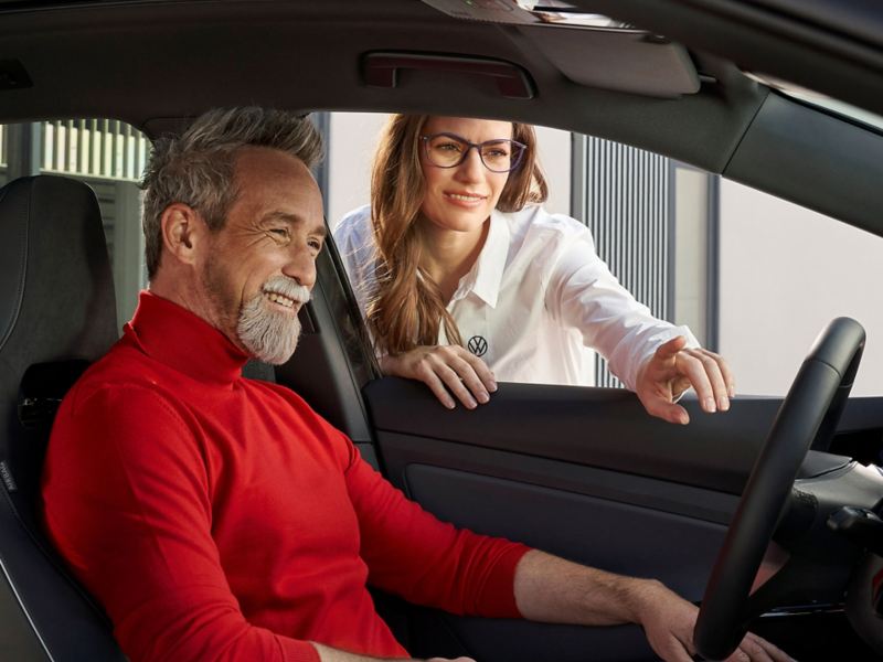 Person im roten Pullover sitzt auf Fahrersitz eines Fahrzeugs mit geöffnetem Fenster und erhält Instruktionen einer VW Mitarbeiterin