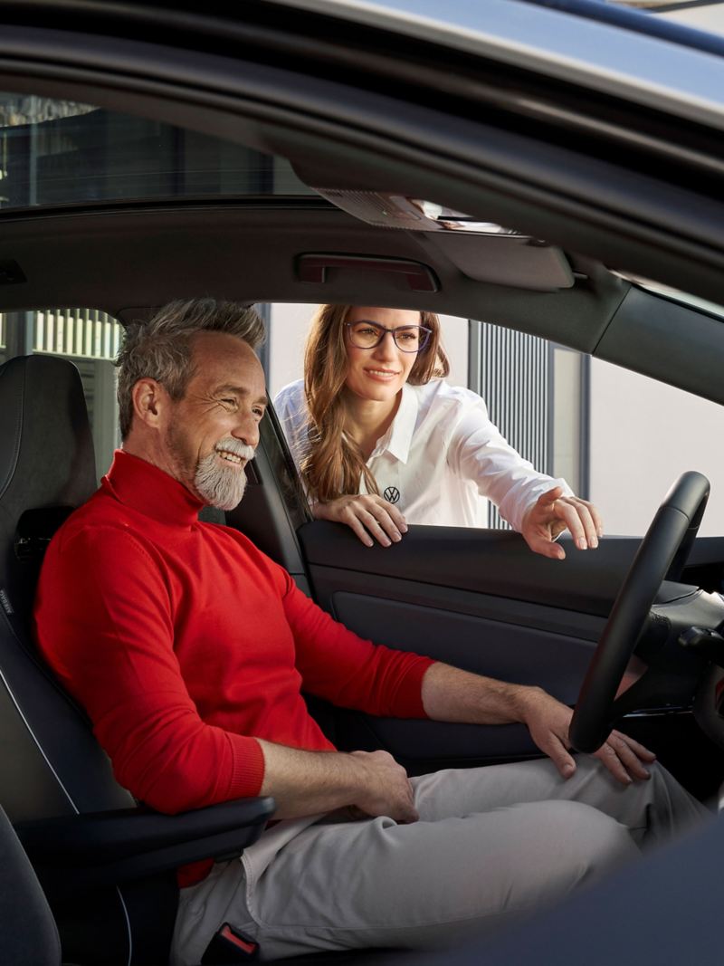Person im roten Pullover sitzt auf Fahrersitz eines Fahrzeugs mit geöffnetem Fenster und erhält Instruktionen einer VW Mitarbeiterin