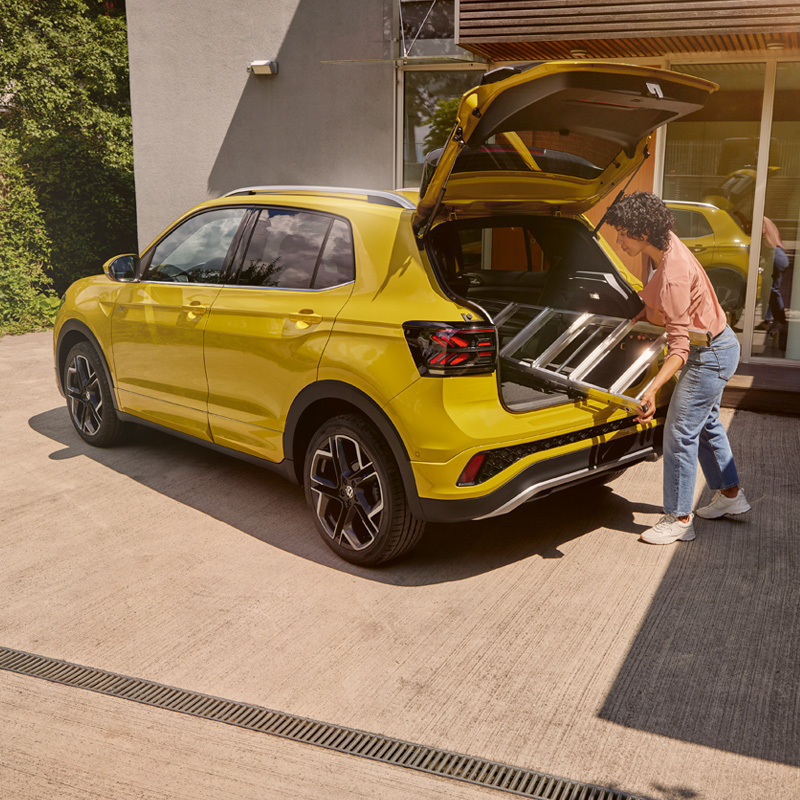 Gele Volkswagen T-Cross met open achterklep: een vrouw laadt een aluminium ladder in de kofferbak.
