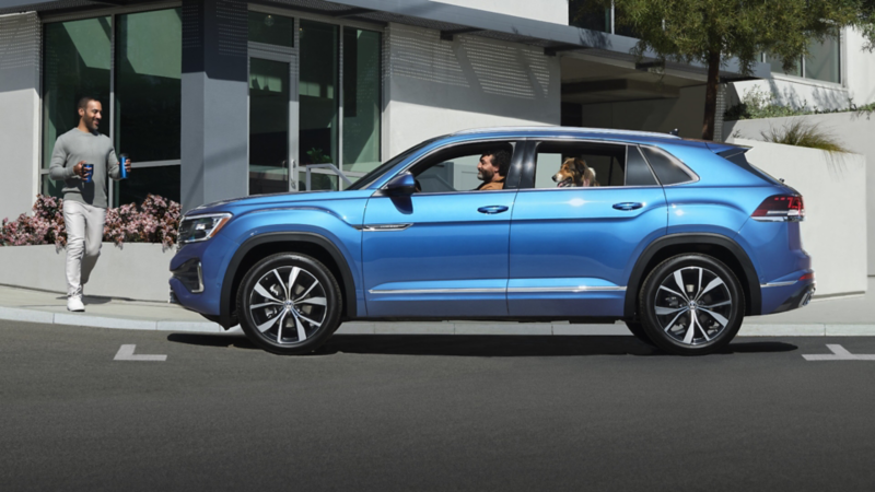 A man with a dog in a blue VW Atlas Cross Sport parked by a building