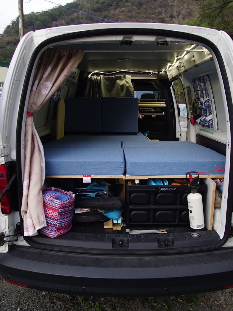 白色Caddy Van後尾門左右開啟，看到車主自行改裝的後廂空間，墊高的木板上平放藍色睡墊，木板下方則有充裕的收納空間置放雜物及生活用品