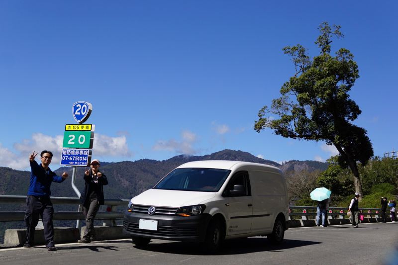 車主與太太在路標旁拍照，旁邊停著白色Caddy Van、後方是藍天白雲及山巒線