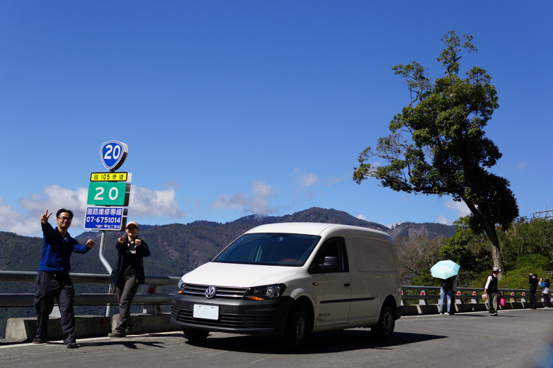 車主與太太在路標旁拍照，旁邊停著香草白色Caddy Van、後方是藍天白雲及山巒線