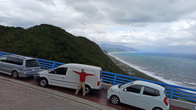 白色Caddy Van停在可看見海洋的橋邊，車主站在車側前方,，雙手往左右平舉比讚