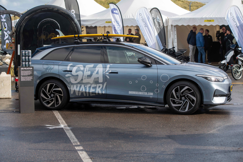Een Volkswagen ID.7 met SEA Water bestickering op de auto staat geparkeerd naast partytenten. Op de achtergrond zie je duinen