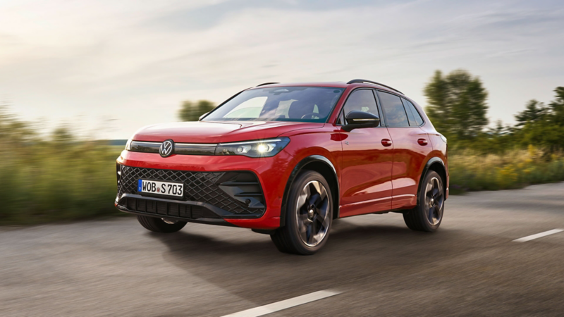 Un SUV Volkswagen rouge roule sur une route de campagne, les phares allumés ; le paysage en arrière-plan apparaît légèrement flou en raison du mouvement.