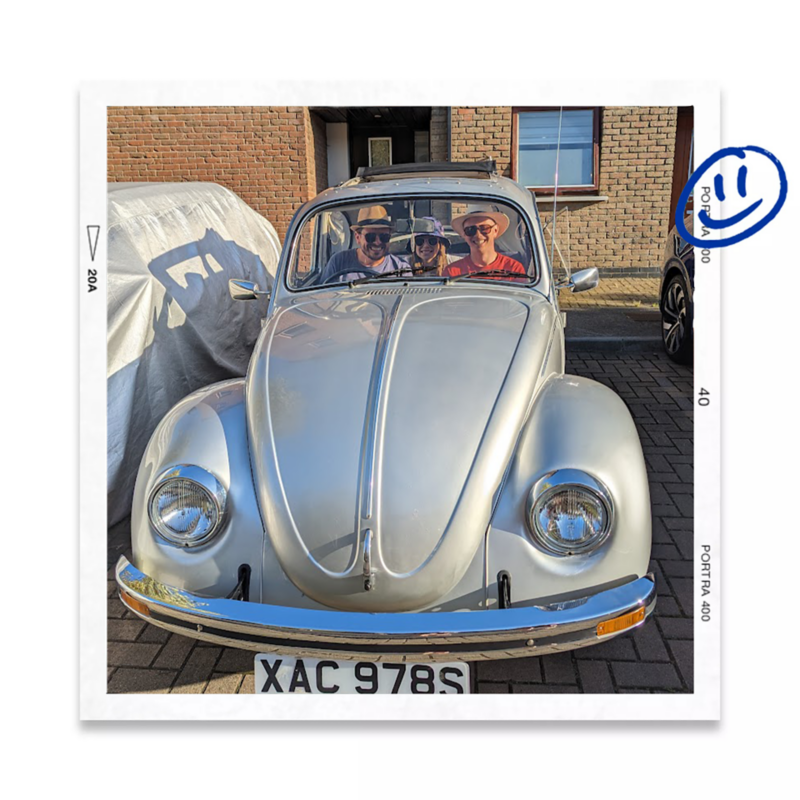 Front view of a classic Volkswagen Beetle parked outdoors with people seated inside the car, smiling through the windscreen