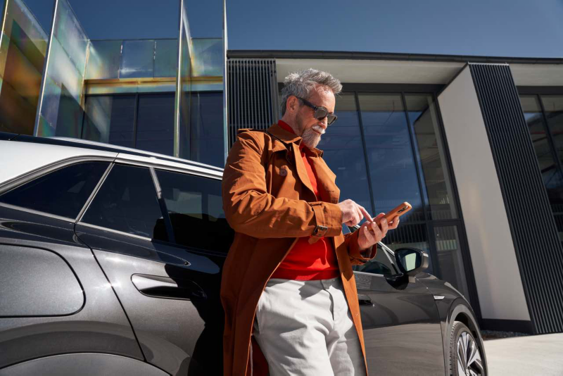 Homme en manteau orange utilisant un smartphone, adossé à une voiture noire devant un bâtiment moderne.