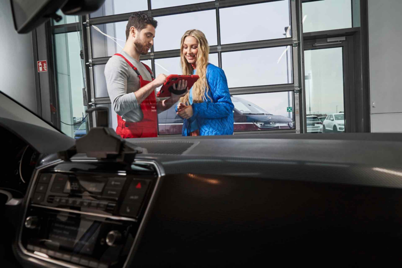 Technicien en salopette rouge consultant une tablette, en discussion avec une cliente dans un atelier automobile.