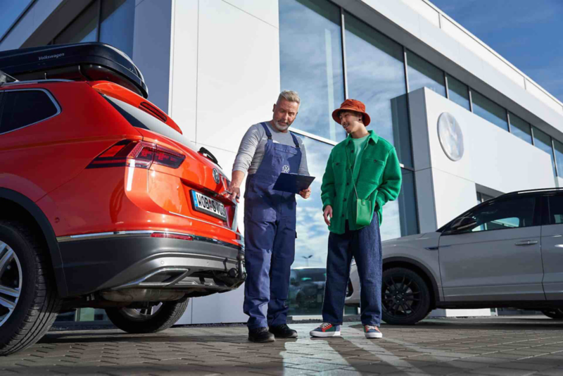 SUV Tiguan orange avec coffre de toit devant concession Volkswagen, deux personnes discutant à côté.
