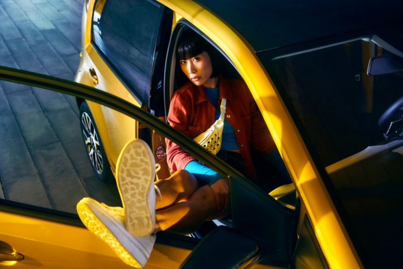 Parkender VW e-up in Gelb mit geöffneter Beifahrertür. Frau auf Beifahrersitz lehnt Füße aus dem Fenster.