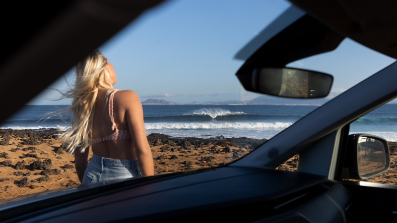 Une femme blonde, vue de dos, regarde les vagues de l'océan depuis un rivage rocheux, le tout encadré par l'intérieur d'un caddy california vw.