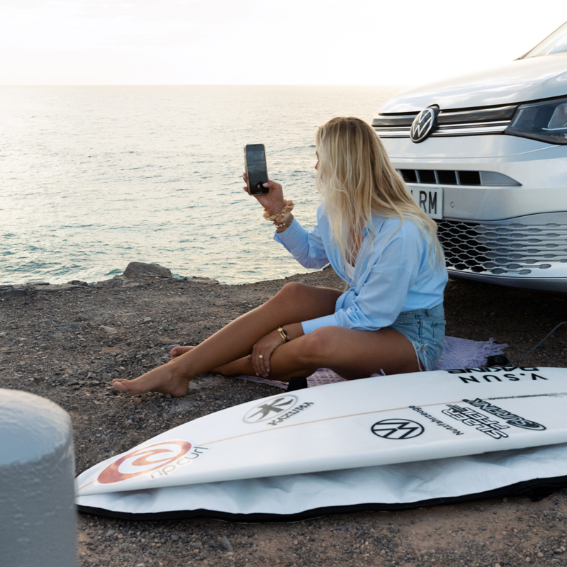 Une femme blonde est assise sur le sol, face à la mer, tenant un téléphone. À ses côtés se trouvent un caddy california vw Volkswagen gris et une planche de surf.
