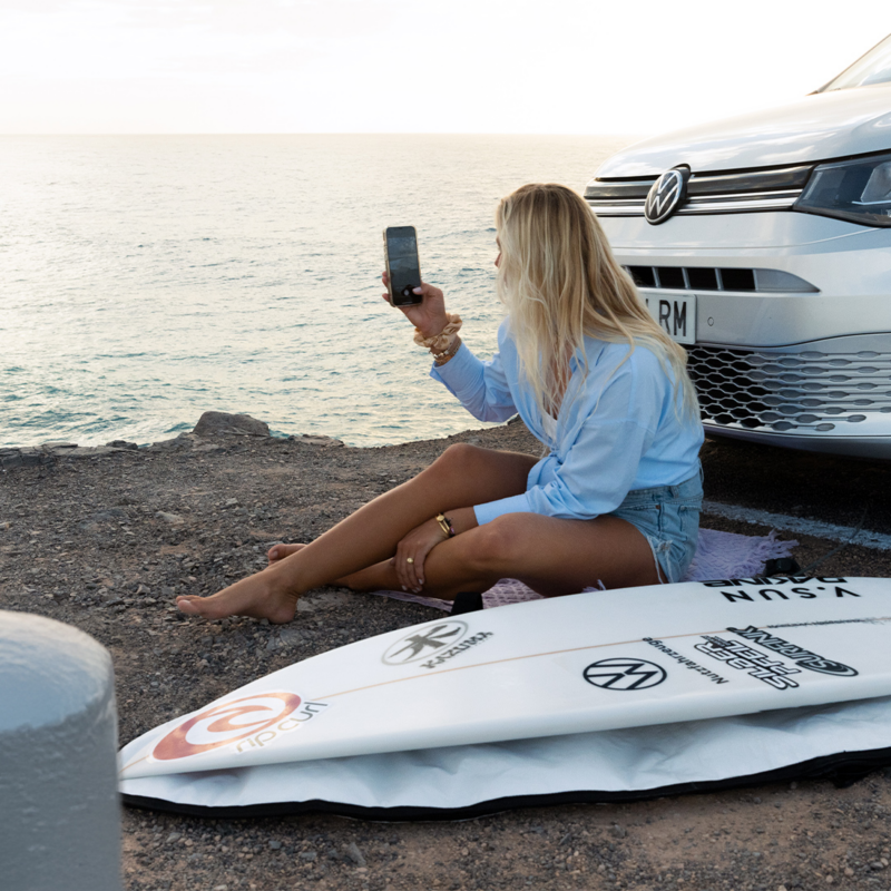 Une femme blonde est assise sur le sol, face à la mer, tenant un téléphone. À ses côtés se trouvent un caddy california vw Volkswagen gris et une planche de surf.