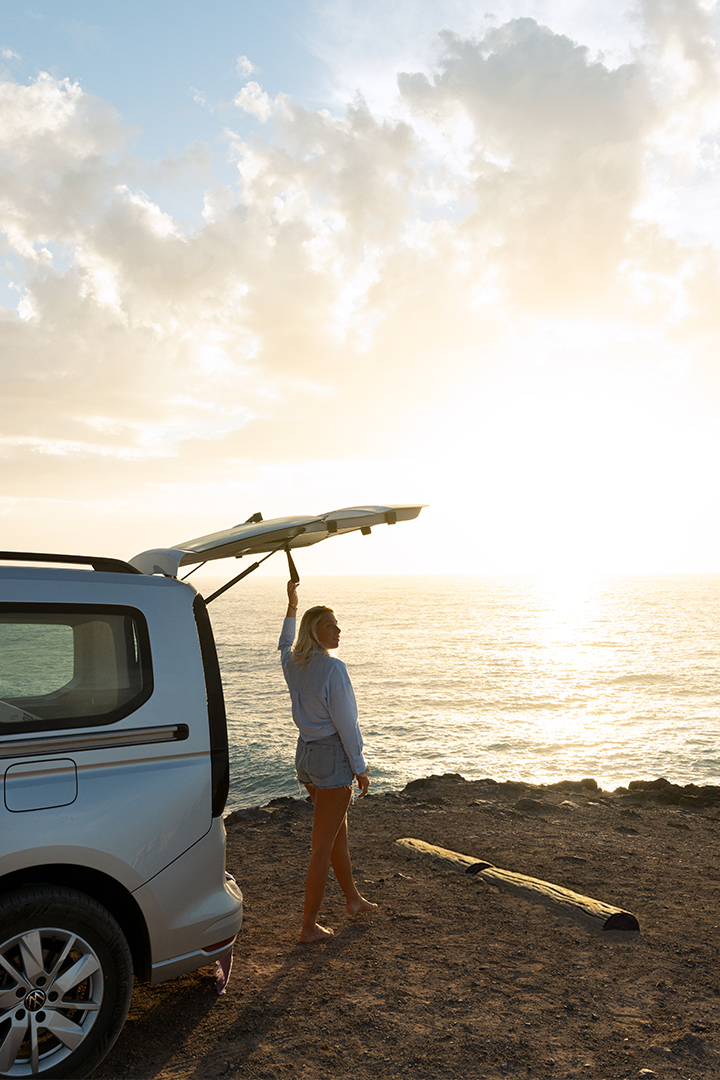 Une femme blonde, pieds nus, se tient au bord d'une falaise, le hayon d'un van Volkswagen VW caddy California clair ouvert, face à l'océan au coucher du soleil.