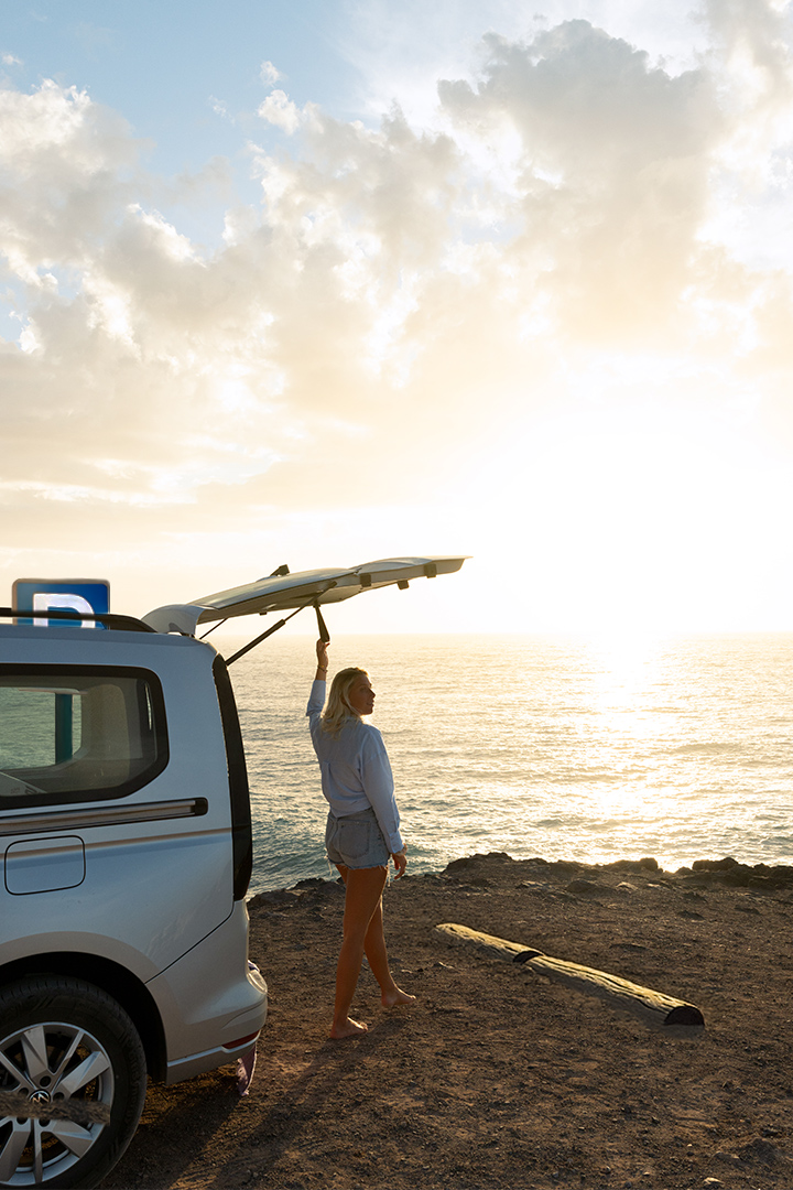 Une femme blonde, pieds nus, se tient au bord d'une falaise, le hayon d'un van Volkswagen VW caddy California clair ouvert, face à l'océan au coucher du soleil.