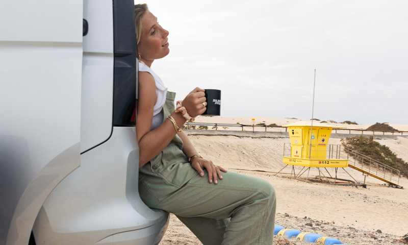 Une femme blonde est assise sur le pare-chocs d'un van vw caddy california, tenant une tasse, face à une plage avec une tour de sauveteur jaune et des dunes de sable.