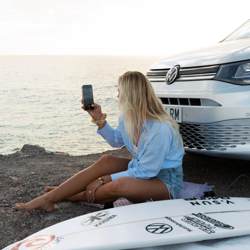 Une femme blonde est assise sur le sol, face à la mer, tenant un téléphone. À ses côtés se trouvent un caddy california vw Volkswagen gris et une planche de surf.