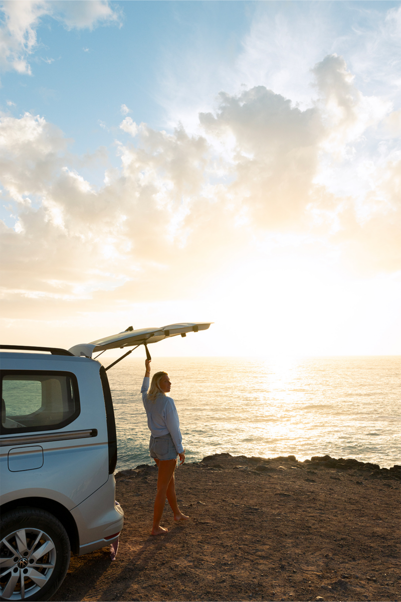 Une femme blonde, pieds nus, se tient au bord d'une falaise, le hayon d'un van Volkswagen VW caddy California clair ouvert, face à l'océan au coucher du soleil.