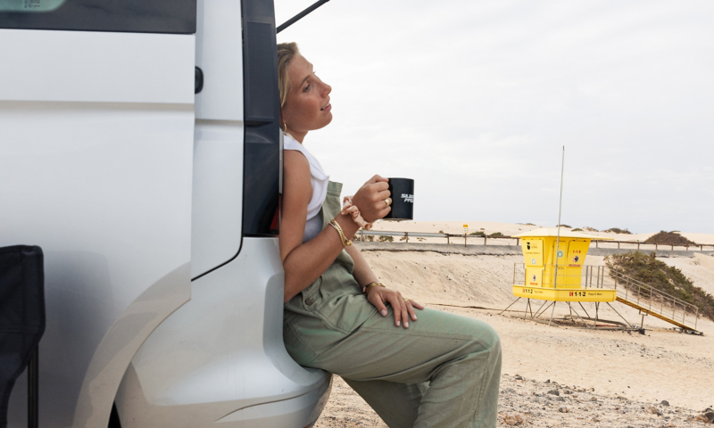 Surferin sitzt an der geöffneten Heckklappe des VW Caddy und trinkt einen Kaffee.