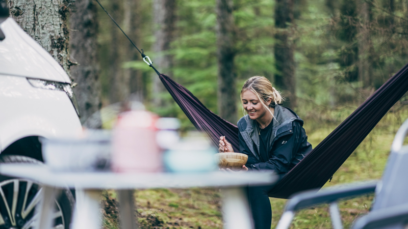 Eine Frau sitzt in einer Hängematte im Wald und isst etwas aus einer Schale.