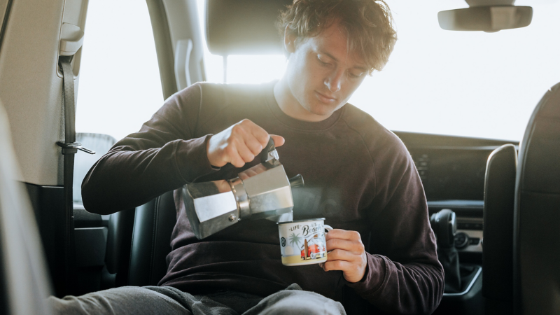 Ein Mann schenkt sich gerade einen Kaffe ein während er auf dem Rücksitz sitzt.