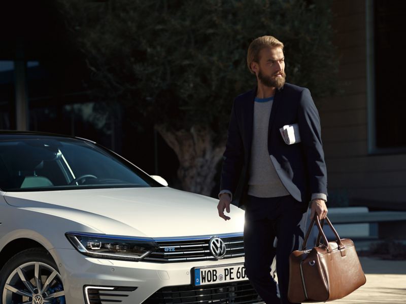 Un uomo con abito elegante e borsa da lavoro passa davanti alla sua Volkswagen Passat GTE per uso aziendale