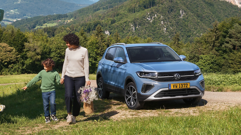Volkswagen T-Cross Life bleu stationné en pleine nature, avec deux personnes marchant à proximité sur un chemin entouré de verdure.