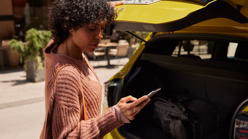 En kvinna står vid bakluckan på en Volkswagen och tittar ner i sin mobiltelefon