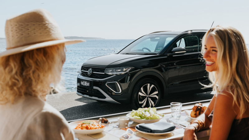 Two women sat in a restaurant with a view over ocean and T-Cross parked up
