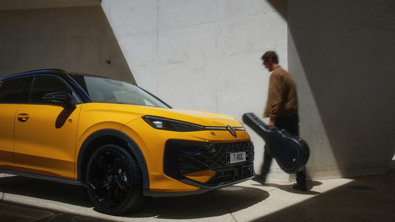 A man with a guitar case walks past a yellow VW T-Roc R-Line in modern architecture.