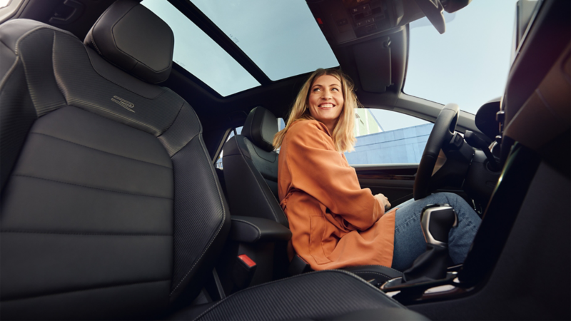 Une femme est assise dans une voiture, sur le siège passager. Elle porte une veste orange et sourit en regardant sur le côté. La voiture a un intérieur en cuir noir. On peut voir le ciel à travers le toit ouvrant.