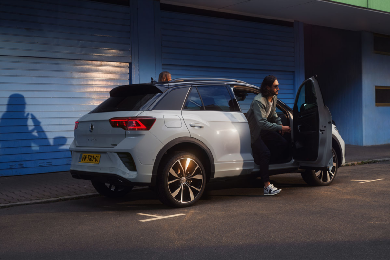 Man leunt tegen een witte Volkswagen T-Roc die geparkeerd staat naast een gebouw