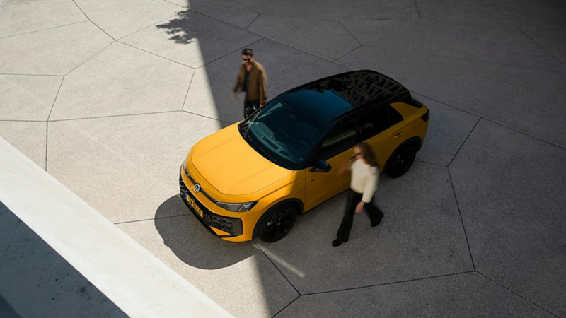 T-Roc jaune photographié à vol d'oiseau. Une femme et un homme se tiennent à côté de la voiture, côté conducteur et côté passager.