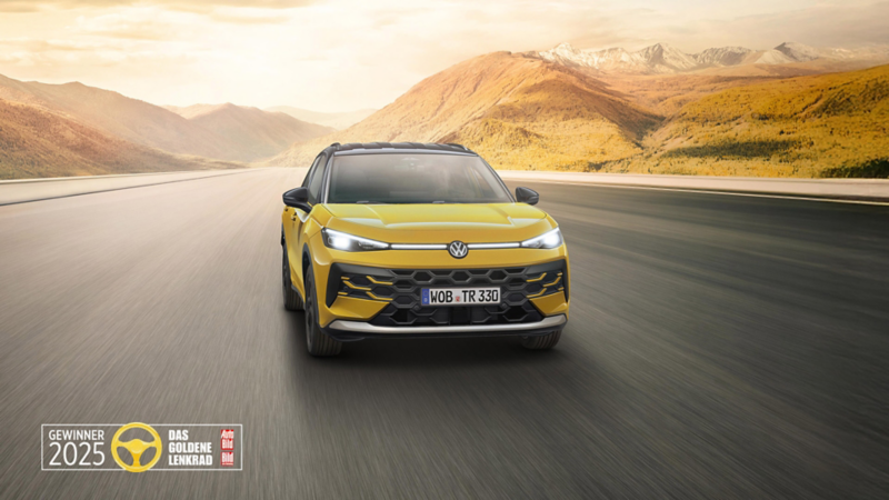 Alttext: Ein gelber VW T-Roc fährt im Sonnenaufgang auf einer Straße durch die Landschaft. Ebenfalls abgebildet ist das Logo des Goldenen Lenkrads 2025.
