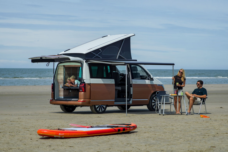 Una furgoneta camper Volkswagen California bicolor (marrón y blanca) con el techo elevable desplegado y toldo extendido, estacionada en una playa de arena junto al mar. Una pareja adulta relaja fuera del vehículo mientras dos niños están dentro. Una tabla de paddle surf naranja y roja descansa en la arena en primer plano, evocando una escena de vacaciones familiares y actividades acuáticas.