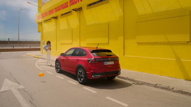 VW Taigo in Rot am Straßenrand vor gelbem Gebäude, Heck- und Seitenansicht, Frau geht auf Fahrzeug zu