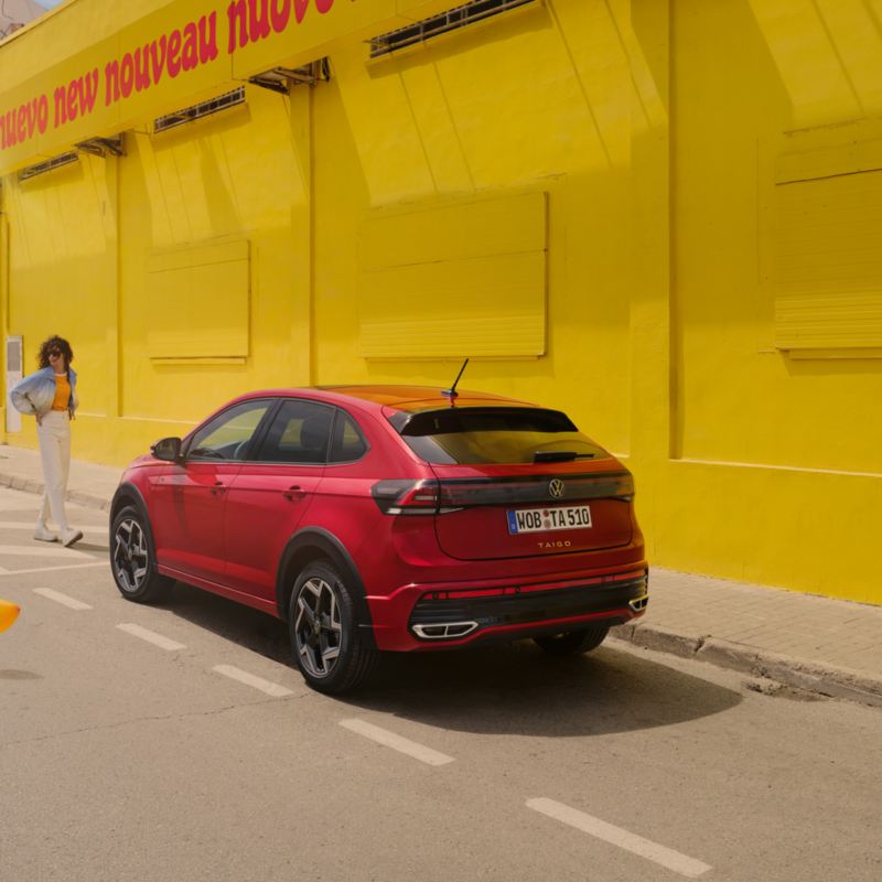 VW Taigo in Rot am Straßenrand vor gelbem Gebäude, Heck- und Seitenansicht, Frau geht auf Fahrzeug zu